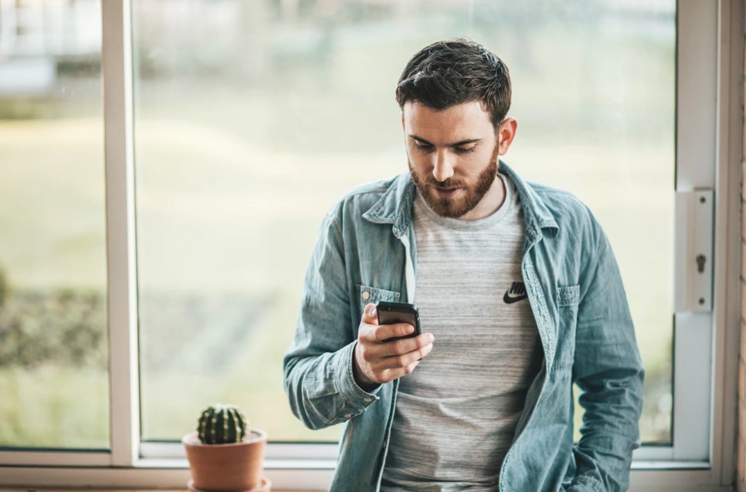 Man leaning against window looking and holding smartphone.