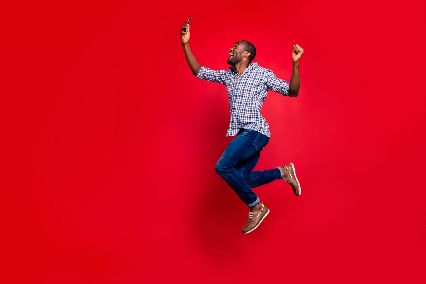 Photo of a young man of African descent jumping in the air while holding and looking at a cell phone. He is smiling and visibly happy. The backdrop is red.